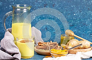 Organic turmeric milk. Composition of glass and decanter with golden milk, ingredients and honey on blue background.