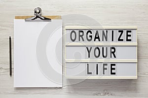 `Organize your life` on a lightbox, clipboard with blank sheet of paper on a white wooden background, top view. Flat lay, overhe