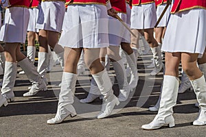 Organized group of girls in bright ceremonial costumes march along a city street with drumsticks in their hands.