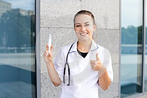 Oriental smiling female nurse with stethoscope in medical gown holding syringe up and pointing thumbs up