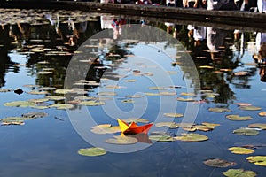 Origami boat in a pond with lilies on the water and reflections of people and trees. Beautiful landscape with paper boat