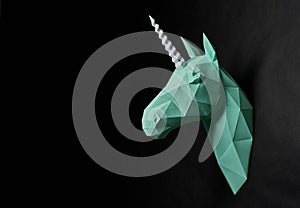 Origami dull green unicorn`s head hanging on black wall.