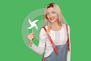Origami hand mill. Portrait of beautiful adult blond woman in stylish overalls holding paper windmill
