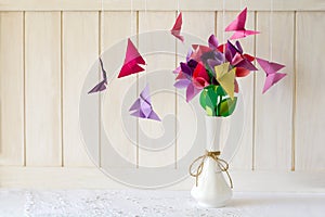 Origami paper flowers in vase and butterflies on white wooden wall.