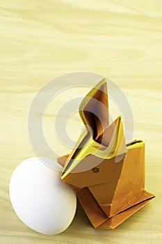 Origami rabbit from orange colored paper. Easter bunny and white chicken egg on a background of a light yellow wooden table, copy