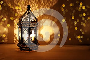 Ornamental Arabic lantern with burning candle glowing at night and glittering golden bokeh lights. Festive greeting card