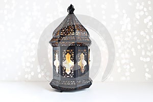 Ornamental dark Moroccan, Arabic lantern on the white table. Burning candle, glittering bokeh lights. Greeting card for