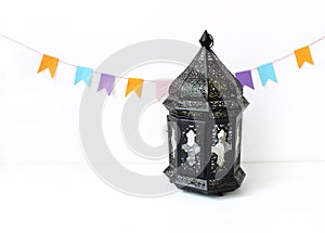 Ornamental dark Moroccan, Arabic lantern on the white table. Party decoration, string of colorful paper flags. Greeting