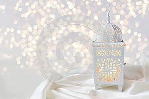 Ornamental white Moroccan, Arabic lantern on silk textile throw. Burning candle, glittering bokeh lights. Greeting card