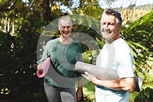 ortrait of happy senior caucasian couple holding yoga mats, looking to camera in sunny garden