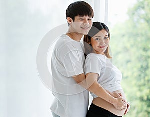 Ortrait of a young Asian couple man and pregnant woman dressed casually smiling and standing together by the window with a warm