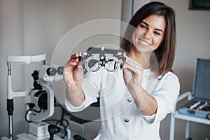 Other equipment in the room. Young attractive female ophthalmologist with special device for testing eyes standing in