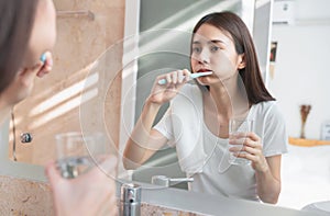 oung Asian woman brushing teeth and looking in the mirror, holding water glass, towel on the shoulder on bathroom.