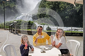 Oung happy family mom, dad and daughter are drinking coffee and eating ice cream at a table in a cafe on the background of a water