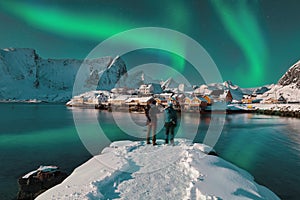 Ð¡ouple of tourists admire the view of the Sakrisoy village and snowy mountaines on background with Northern Lights