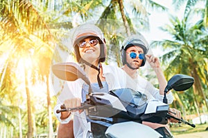 Ð¡ouple travelers riding motorbike scooter in safety helmets during tropical vacation under palm trees