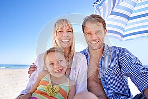 Our holiday house is just off the coast. a happy young family sitting together under a beach umbrella.