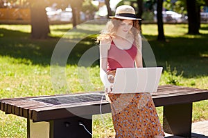 Out door portrait of attrective girlwith foxy hair working with laptop and using wireless Internet, charging her device via solar