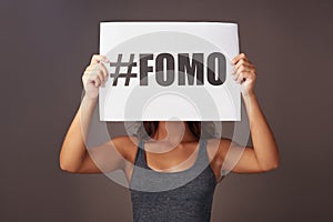 Out of sight, never out of mind. Studio shot of a young woman holding a sign with FOMO printed on it against a gray