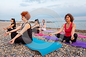 Outdoor activity. Side view of group of Caucasian women meditate sitting on sports mats on pebble beach. Yoga and