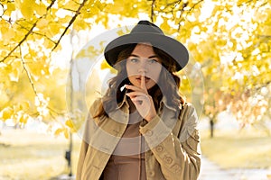Outdoor atmospheric lifestyle portrait of young beautiful lady showing silence gesture. Cute girl in good mood in autumn