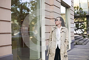 Outdoor autumn portrait of young woman wearing glasses and light color raincoat walking on street