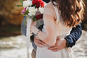 Outdoor beach wedding ceremony near the ocean, close up of embrace of stylish couple with wedding bouquet, man is hugging bride