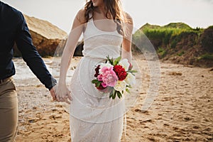 Outdoor beach wedding ceremony near the ocean, close up of hands of stylish couple with wedding bouquet, bride is