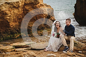 Outdoor beach wedding ceremony near the sea, stylish happy smiling groom and bride are sitting on stones, having fun and