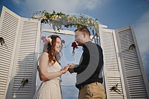 Outdoor beach wedding ceremony, stylish happy smiling groom and bride are standing near wedding altar on the sea shore holding han