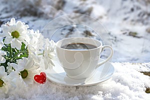 Outdoor breakfast in winter with love, steaming coffee cup, white flowers and a red glass heart on a snow covered garden table on