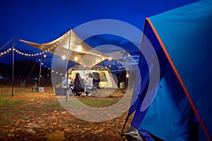 Outdoor camping tent in the forest park, party dinner with friends under tarp or flysheet and warm lighting at night near natural