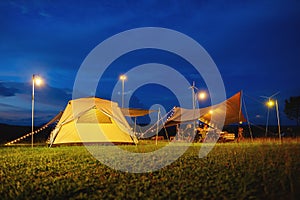 Outdoor camping tent with tarp or flysheet on grass courtyard and warm night light under dark blue sky twilight time, family