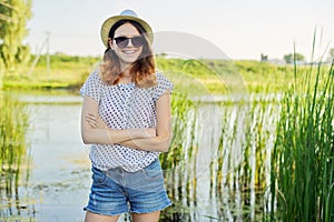 Outdoor country portrait of teenage girl in hat sunglasses near pond in the reeds