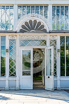 Outdoor facade front view of an old white ornamented glass greenhouse entrance with open door.