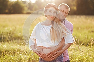 Outdoor family portrait: beautiful young pregnant female and her husband, embrace, keep hands on belly, anticipate child, have wal