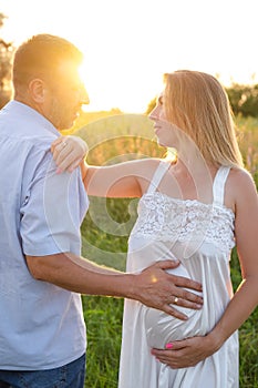 Outdoor family portrait: beautiful young pregnant female and her husband, hands on belly, anticipate child, enjoy