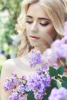 Outdoor fashion beautiful young woman surrounded by lilac flowers summer. Spring blossom lilac bush. Portrait of a girl blond