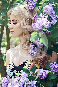 Outdoor fashion beautiful young woman surrounded by lilac flowers summer. Spring blossom lilac bush. Portrait of a girl blond