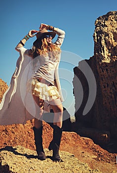 Outdoor fashion photo of young happy woman in hat, sitting on a fur against rocks, hands up