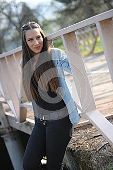 Outdoor fashion portrait of young beautiful fashionable girl with blue shirt and black jeans, standing by the bridge