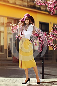 Outdoor full body portrait of young beautiful fashionable happy lady posing near flowering tree. Model wearing stylish