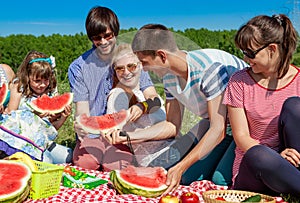 Outdoor group portrait of happy company having picnic on green grass in park and enjoying watermelon