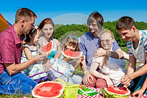 Outdoor group portrait of happy company having picnic on green grass in park and enjoying watermelon