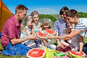 Outdoor group portrait of happy company having picnic on green grass in park and enjoying watermelon