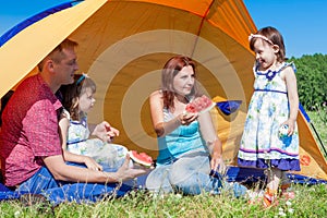 Outdoor group portrait of happy company having picnic near the tent in park and enjoying watermelon