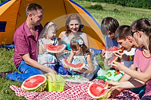 Outdoor group portrait of happy company having picnic near the tent in park and enjoying watermelon