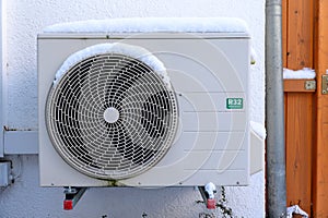 Outdoor heat pump unit covered with snow and R32 refrigerant label sitting on sound and vibration insulating rubber pads against a