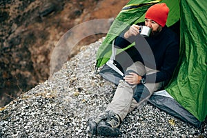 Outdoor horizontal image of young breaded man drinking hot beverage in mountains.