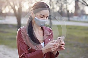 Outdoor image of focused young brunette holding smartphone, using device, wearing protective mask, checking social networking